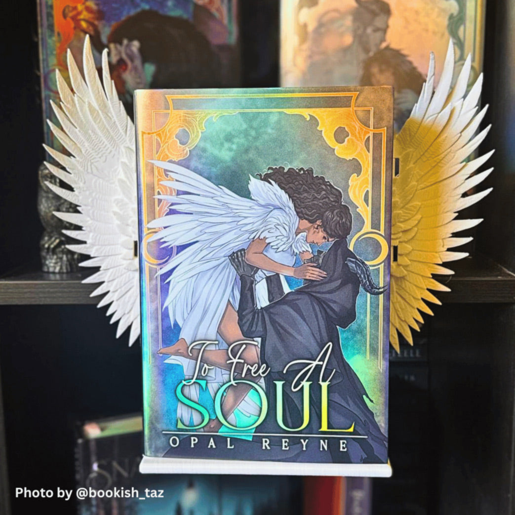 Front-facing view of the fantasy book 'To Free A Soul' by Opal Reyne mounted on a white Curio Candy floating book display hanger with Curio Candy white clip-on angel wings attached to the sides, set on a dark wooden shelf.