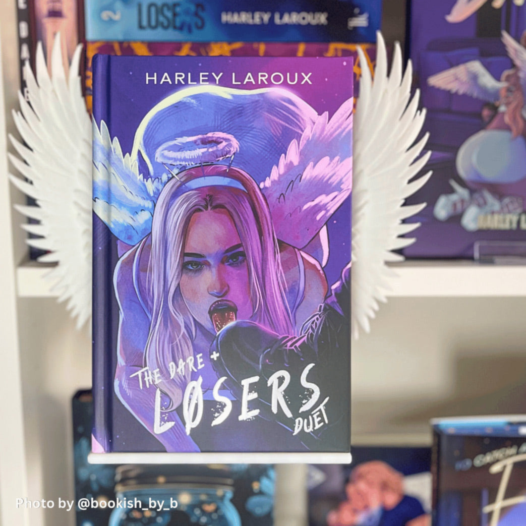 Front-facing view of the dark romance book 'The Dare + Losers Duet' by Harley Laroux mounted on a white Curio Candy floating book display hanger with Curio Candy white clip-on angel wings attached to the sides, set against a bookish shelf display.