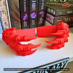 Close-up view of a pair of red 3D printed Dragon Claws Curio Clip-Ons for Curio Candy Floating Book Display Hangers, resting on a bookshelf in front of fantasy romance novels.